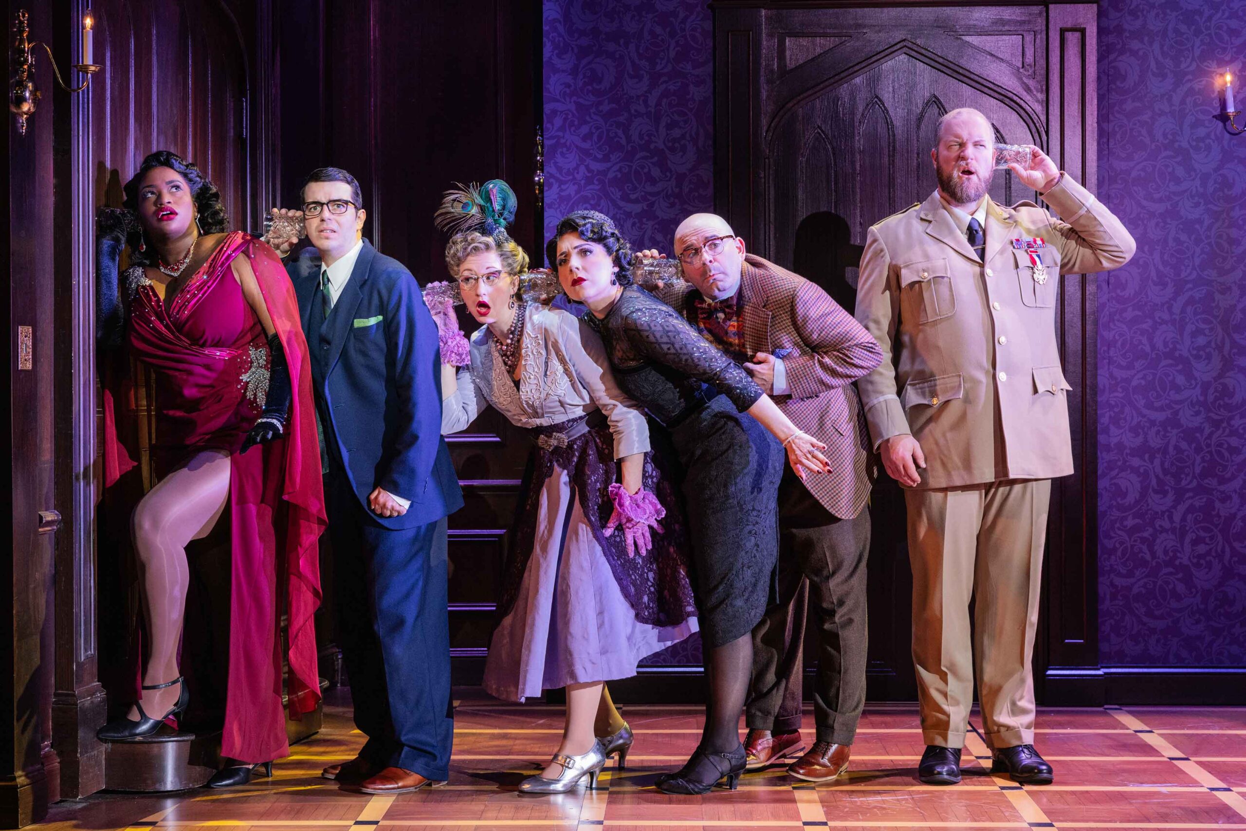 Six actors stand on stage in a line on one side of a closed wooden door, five of them are crouched close together with their ears pressed against glasses, trying to hear a conversation inside the closed door. The other actor is standing facing away from the rest, his glass pressed to his own ear, but the other end is connected to nothing.