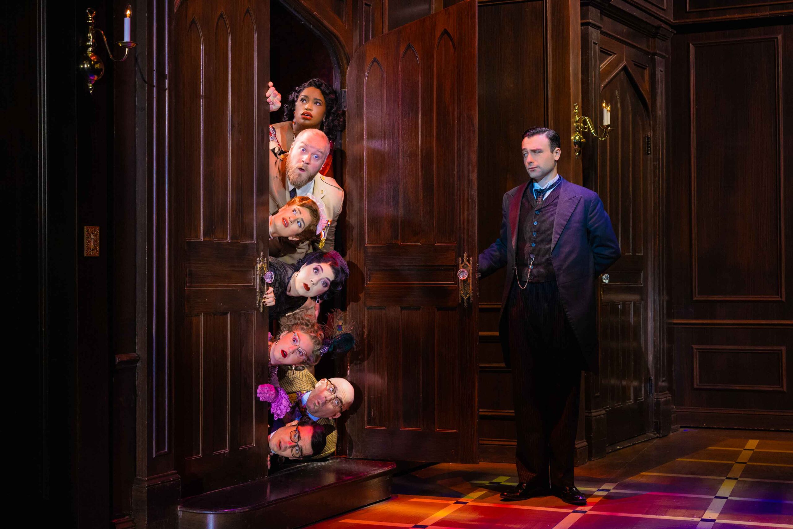 Seven actors lay stacked on top of each other horizontally, peeking out from behind an open door. Only their surprised faces are visible outside of the door frame. Across from them, a man in a suit has one arm outstretched, having just opened the door.